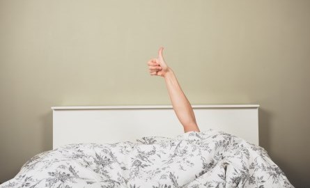 A person in a bed under the covers, with only their arm visible giving a thumbs up